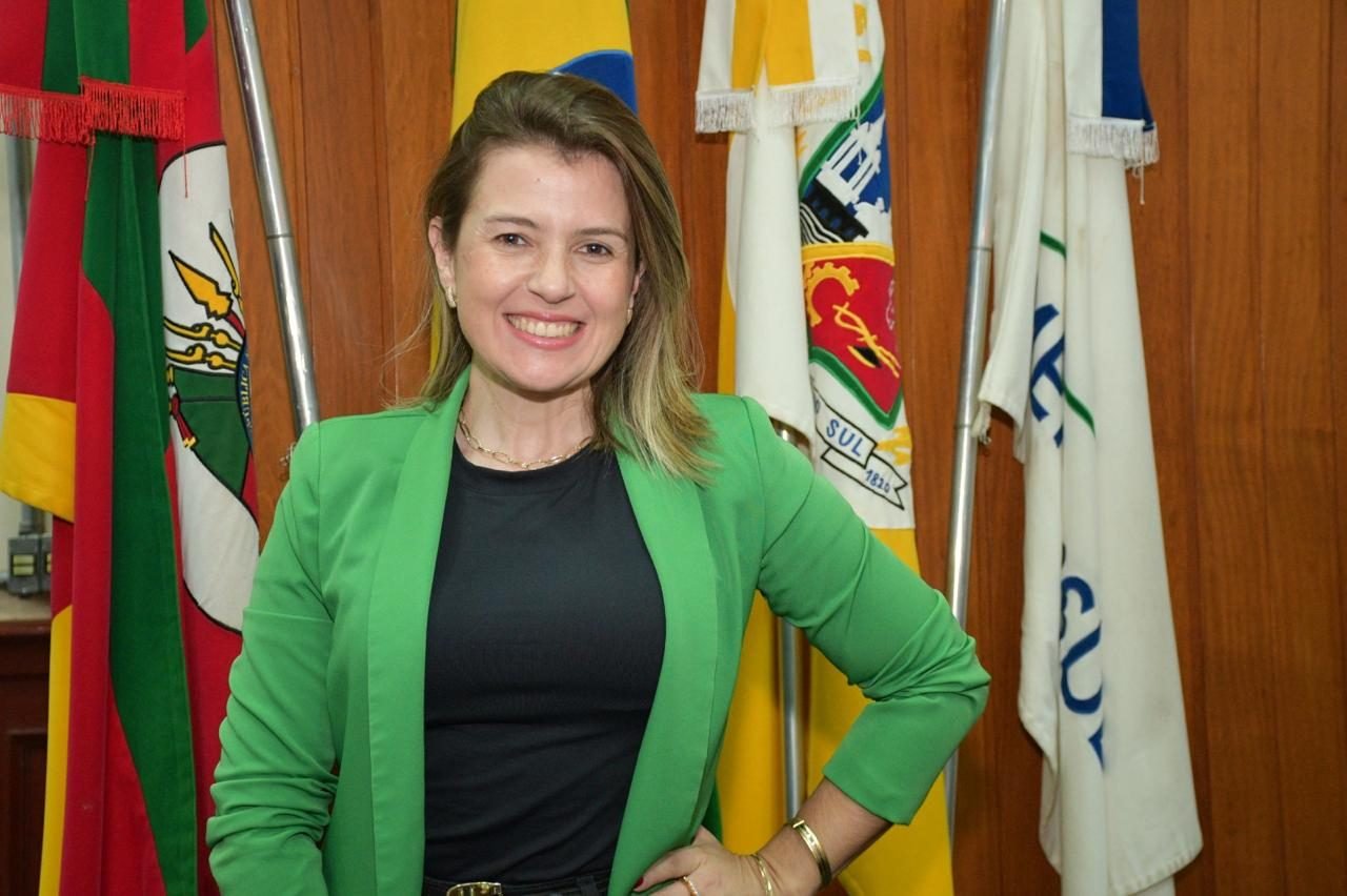 Ana Paula Melo sorri, de blazer verde, diante das bandeiras na Câmara de Vereadores de Cachoeira do Sul